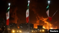 Iran - Iranian flags fly as fire and smoke from an Israeli attack on Sharan Oil depot rise, following Israeli strikes on Iran, in Tehran, June 15, 2025.